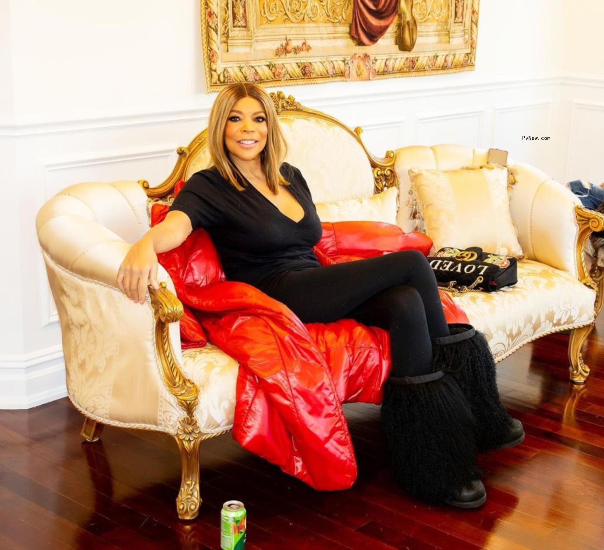 Wendy Williams sitting on couch in black with red coat.