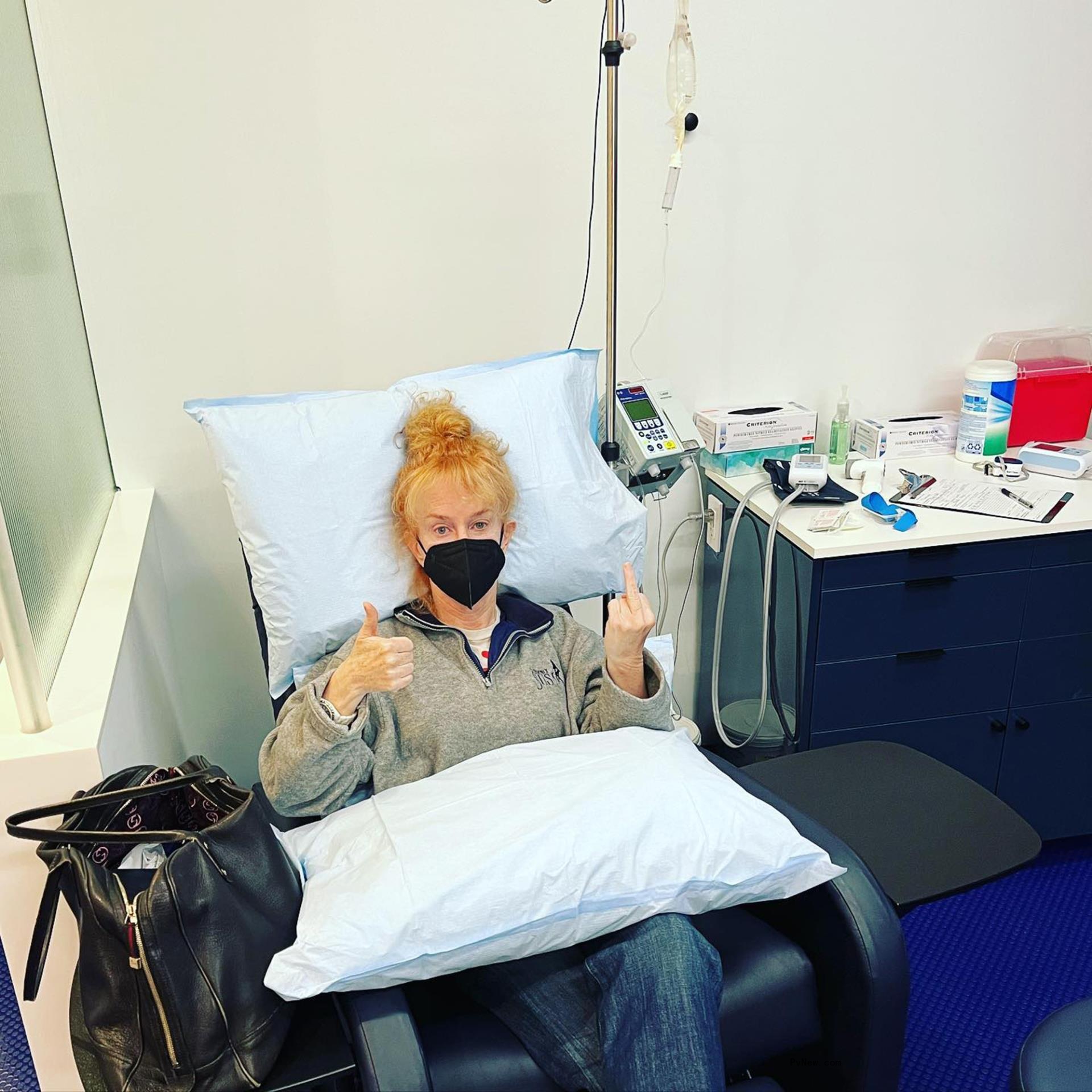 kathy griffin in a mask sitting in a hospital chair with a pillow on her lap