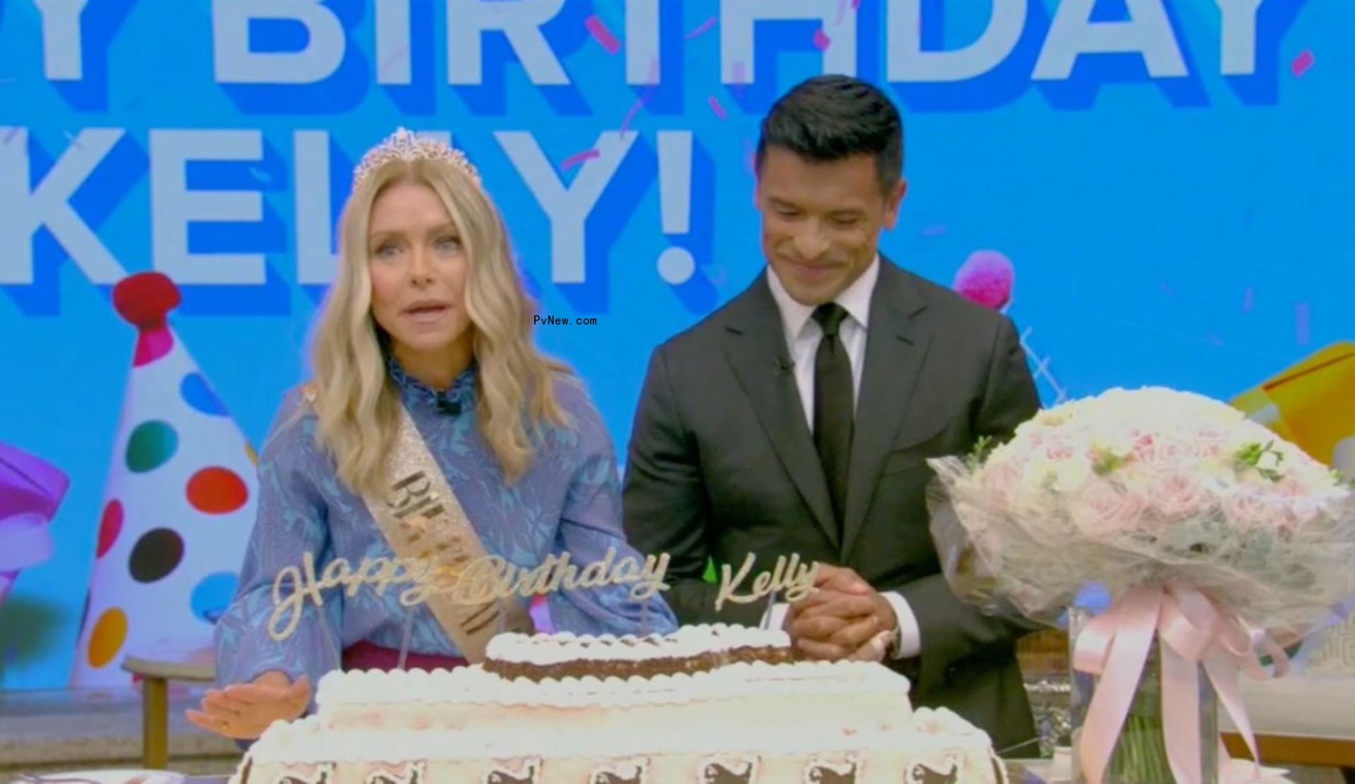 Kelly Ripa in a birthday crown and sash sits next to Mark Co<i></i>nsuelos with a whale-shaped cake in front of her.