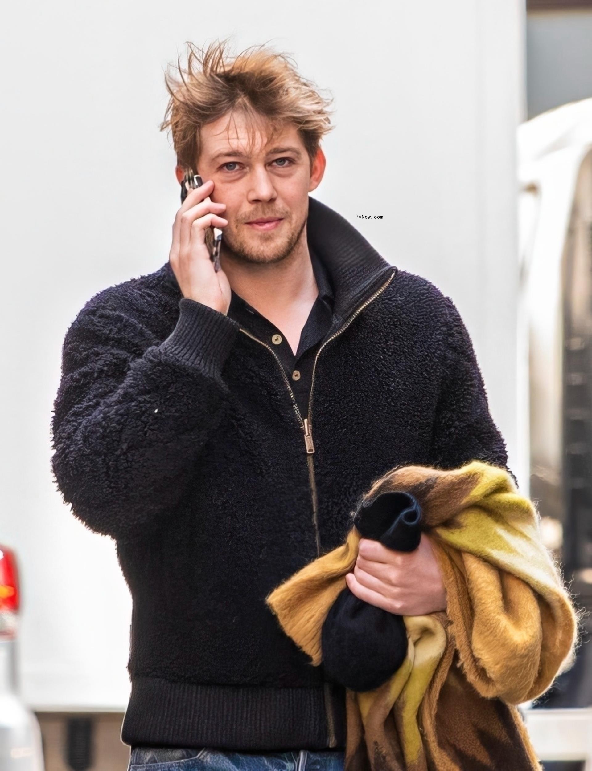 Joe Alwyn walking while holding a cell phone up to his ear.