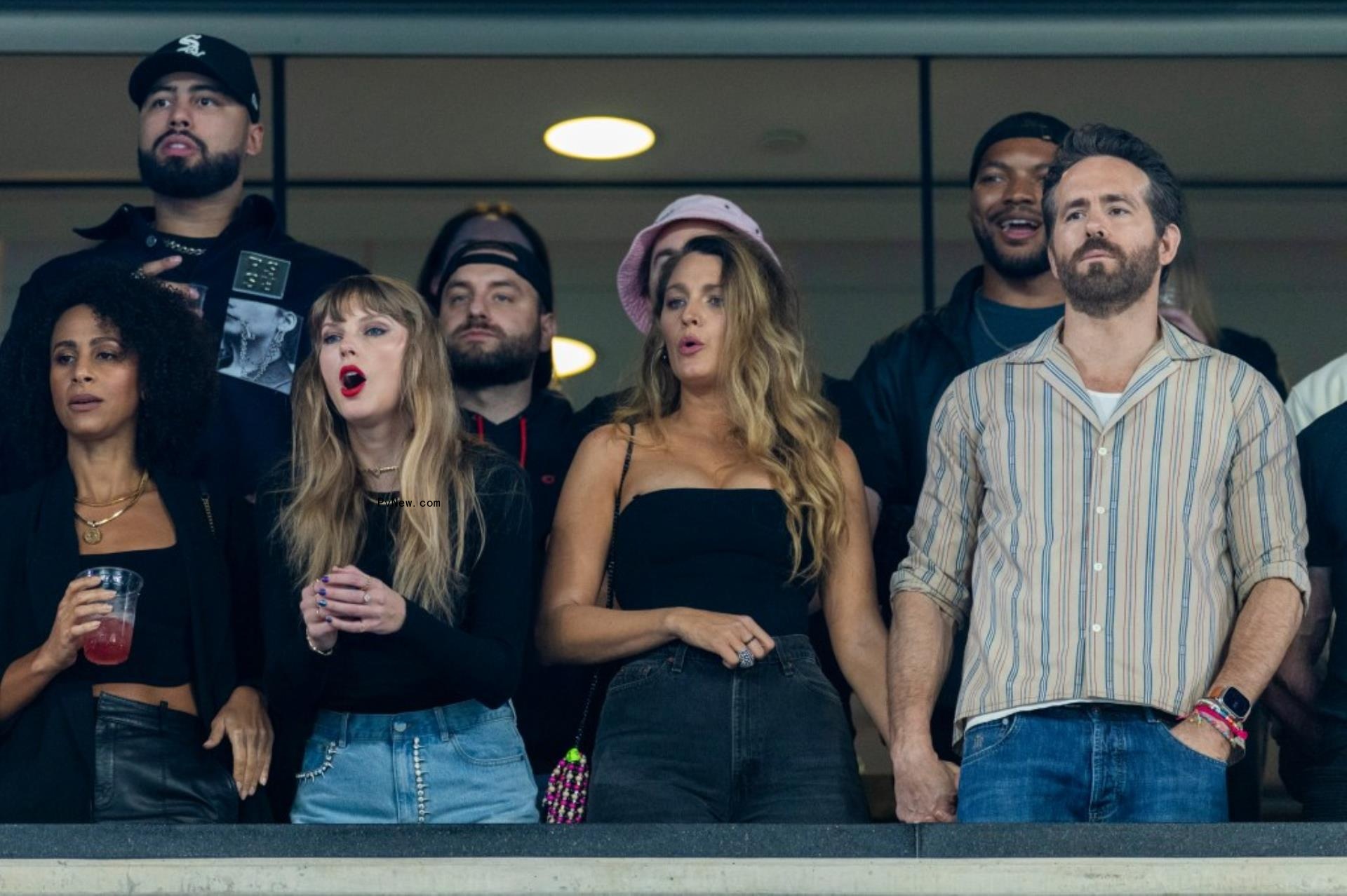 Taylor Swift in black shirt and jean shorts cheering from a VIP booth at a football game