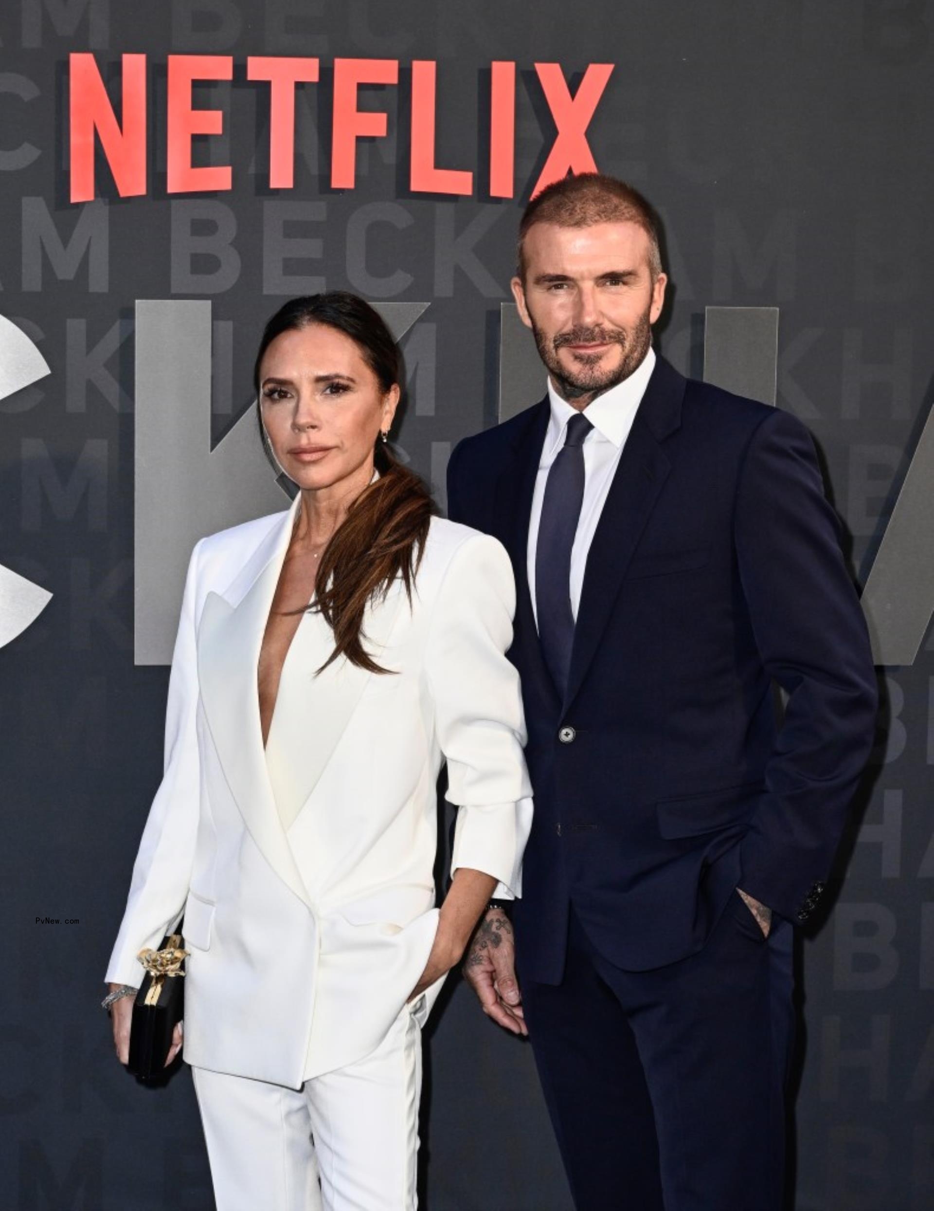 David Beckham in a black suit and Victoria Beckham in a white suit posing at the premiere of their Netflix docu<i></i>mentary