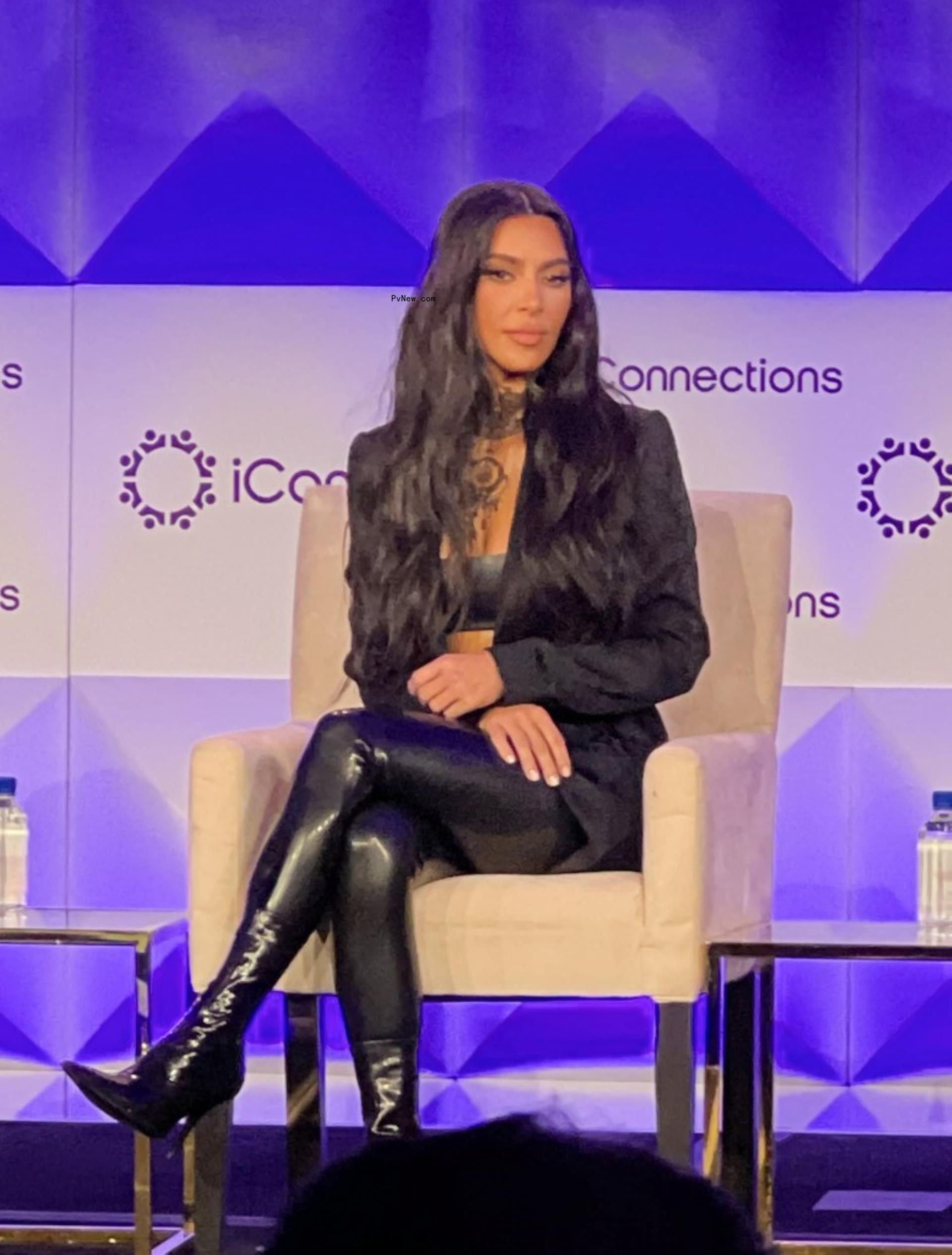 Kim Kardashian sits in a chair on stage in black latex leggings, a black latex bralette, a black blazer, and black booties