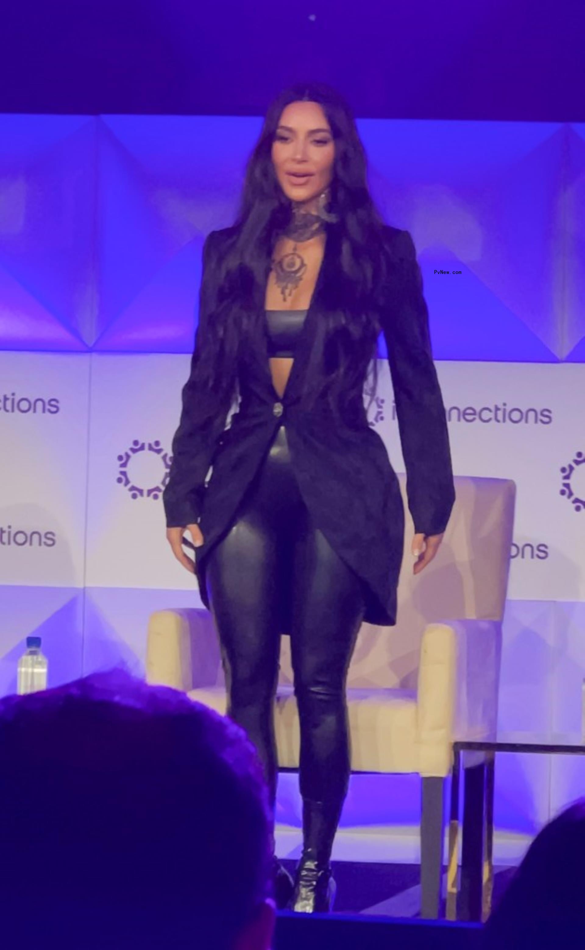 Kim Kardashian stands on stage in black latex leggings, a black latex bralette, a black blazer, and black booties