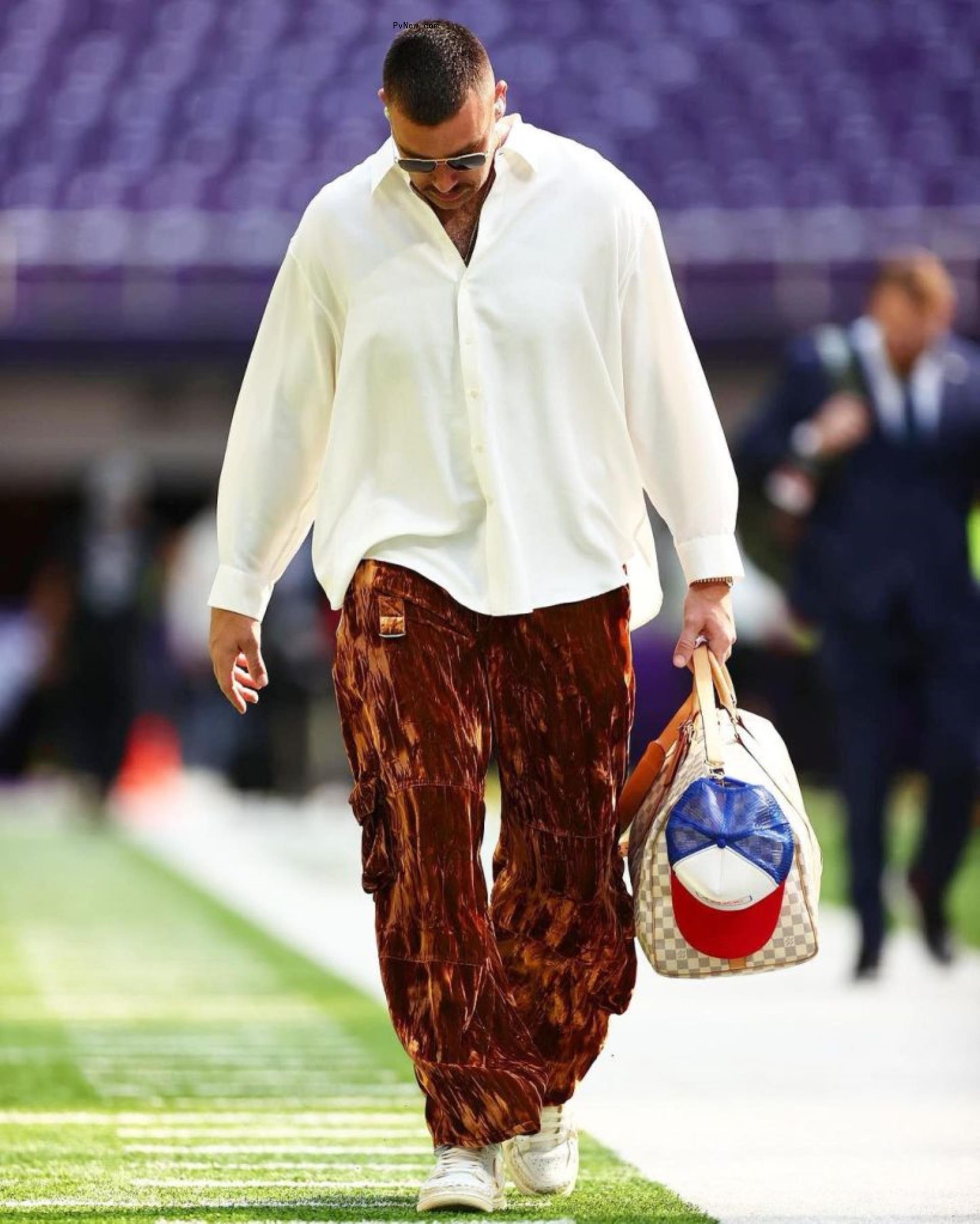 Travis Kelce walking in brown velvety pants that are loose and a flow white button-down shirt. He has a checkered bag in his hands.