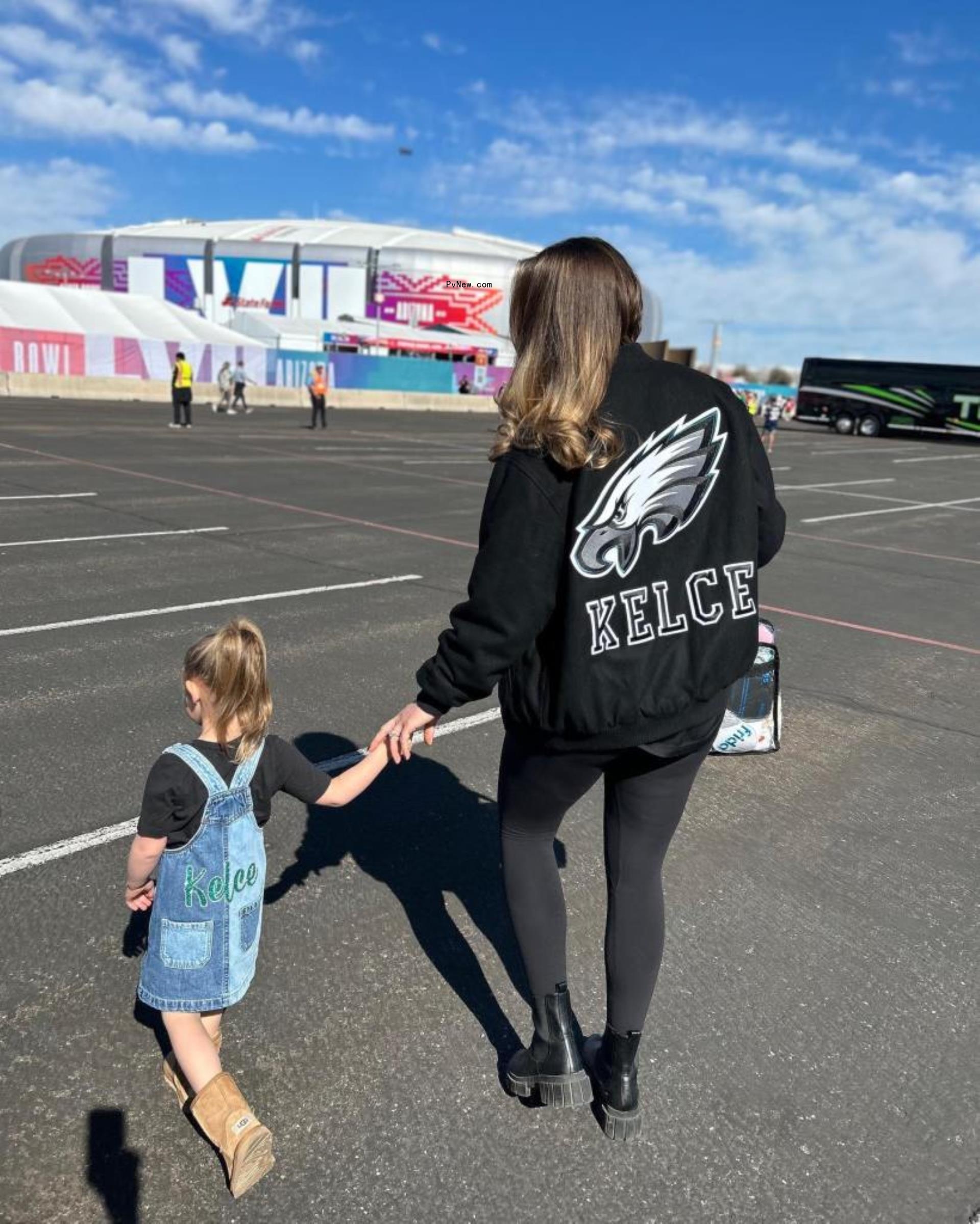Jason Kelce's wife in a black Kelce jacket holds hands with her daughter walking away from camera.