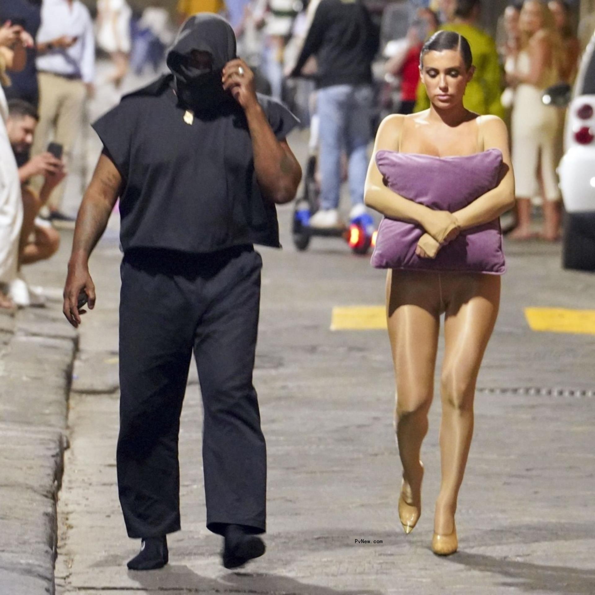 Bianca Censori, holding a pillow to her chest, walking with Kanye West on a street