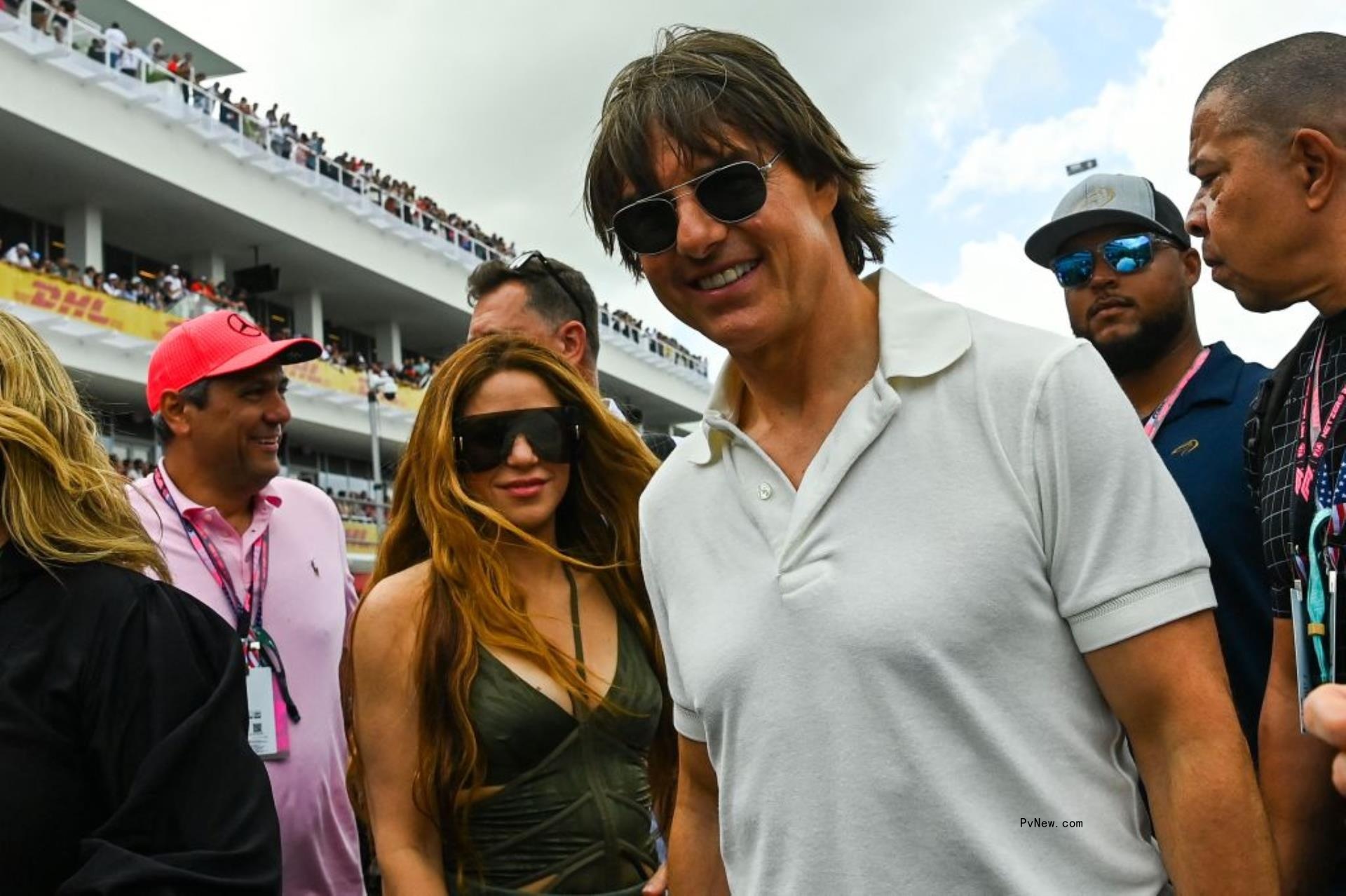 Tom Cruise in a white polo and aviators standing beside Shakira, who is wearing sunglasses and a green low cut tanktop. They are surrounded by men in sunglasses and casual clothes and behind can be seen a grandstand.