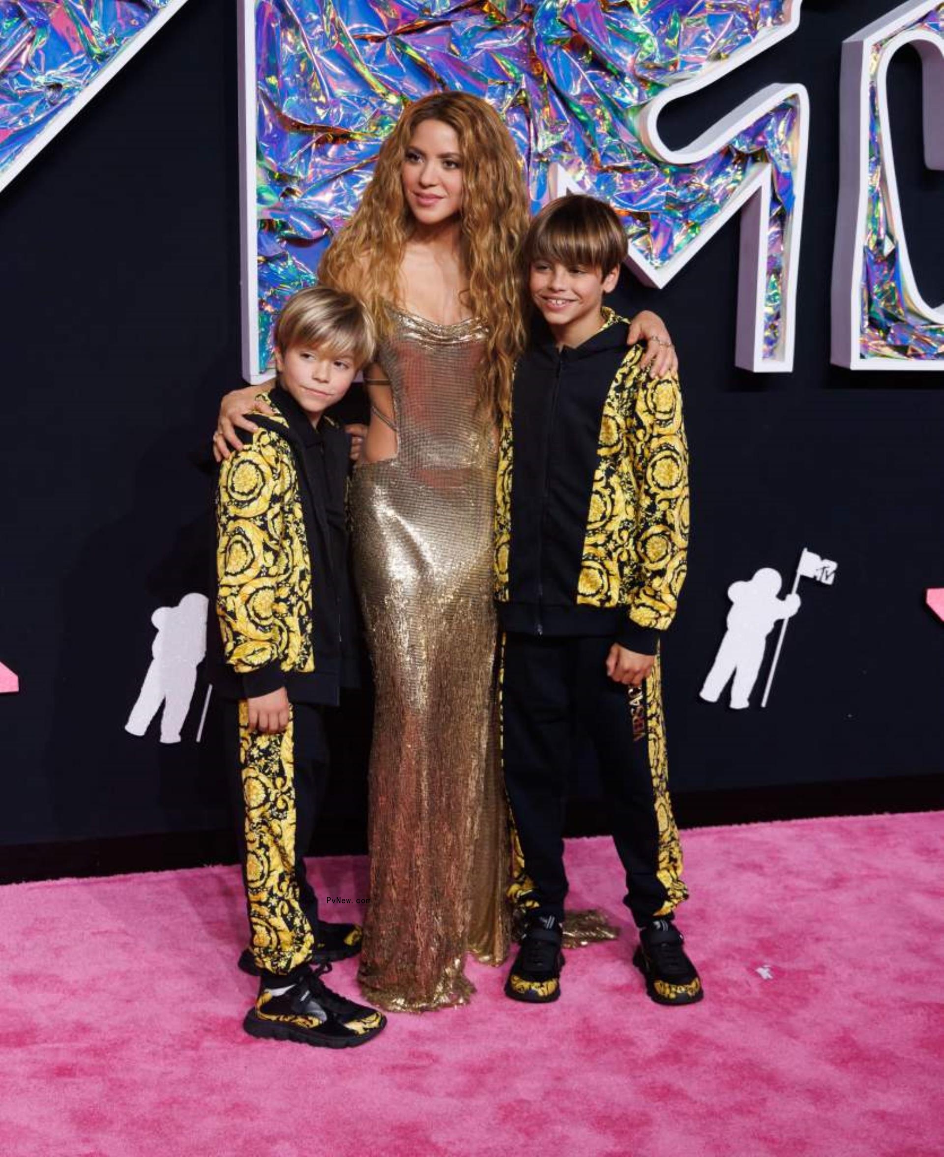 Shakira in a gold sheath dress puts her arms around her sons Mila and Sasha in matching black and yellow track suits on a red carpet.