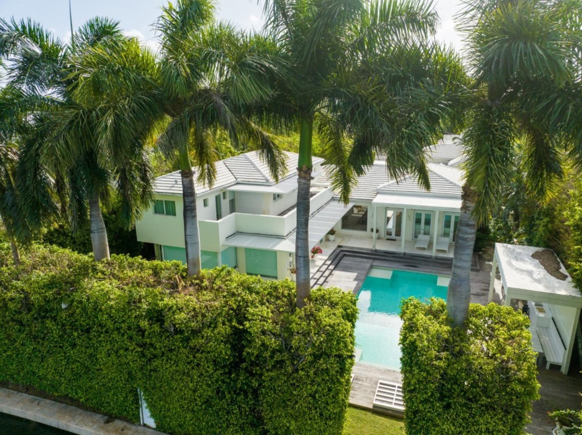 A tropical house seen from behind palm trees and hedges. It has a pool and two stories in white.