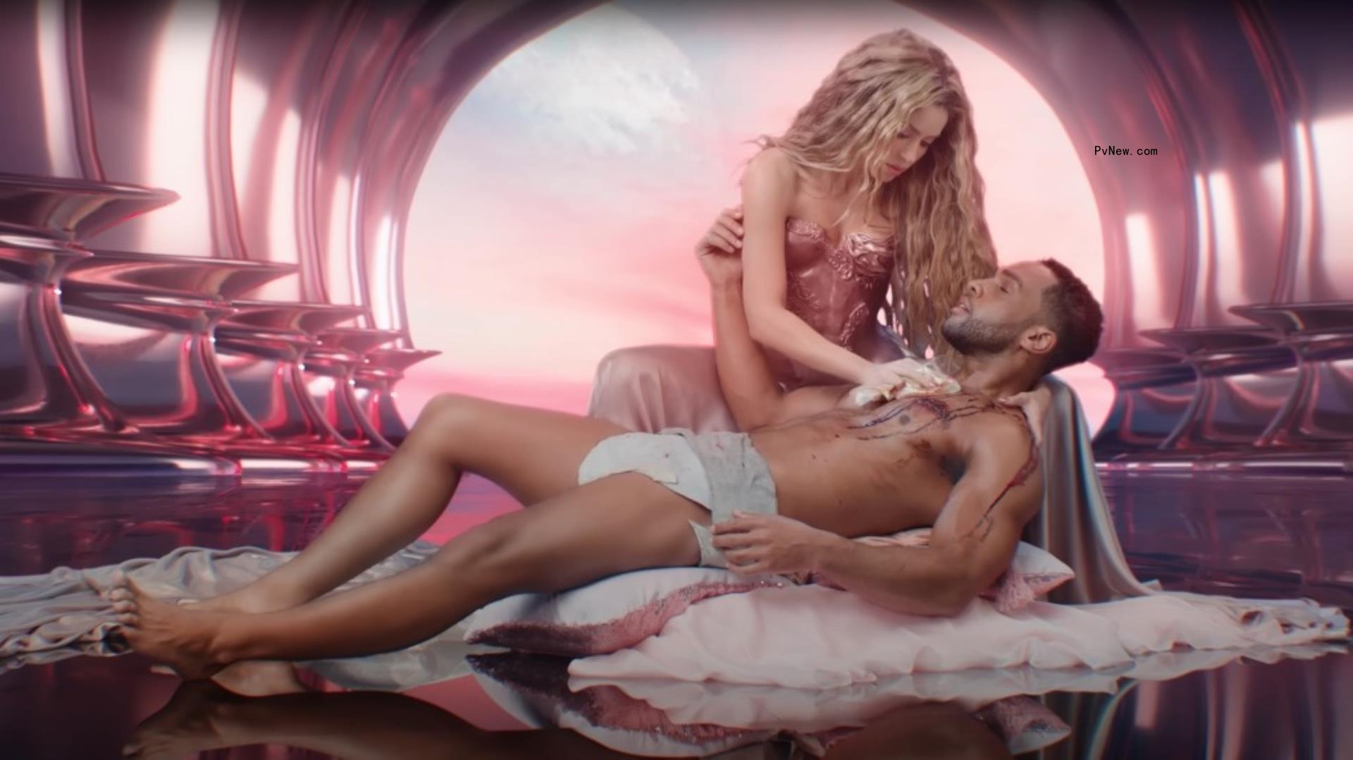 Shakira in a pink bustier leaning over a reclining man who appears to have blood coming from a wound. She is dabbing at it. He appears to be stripped to his underwear.