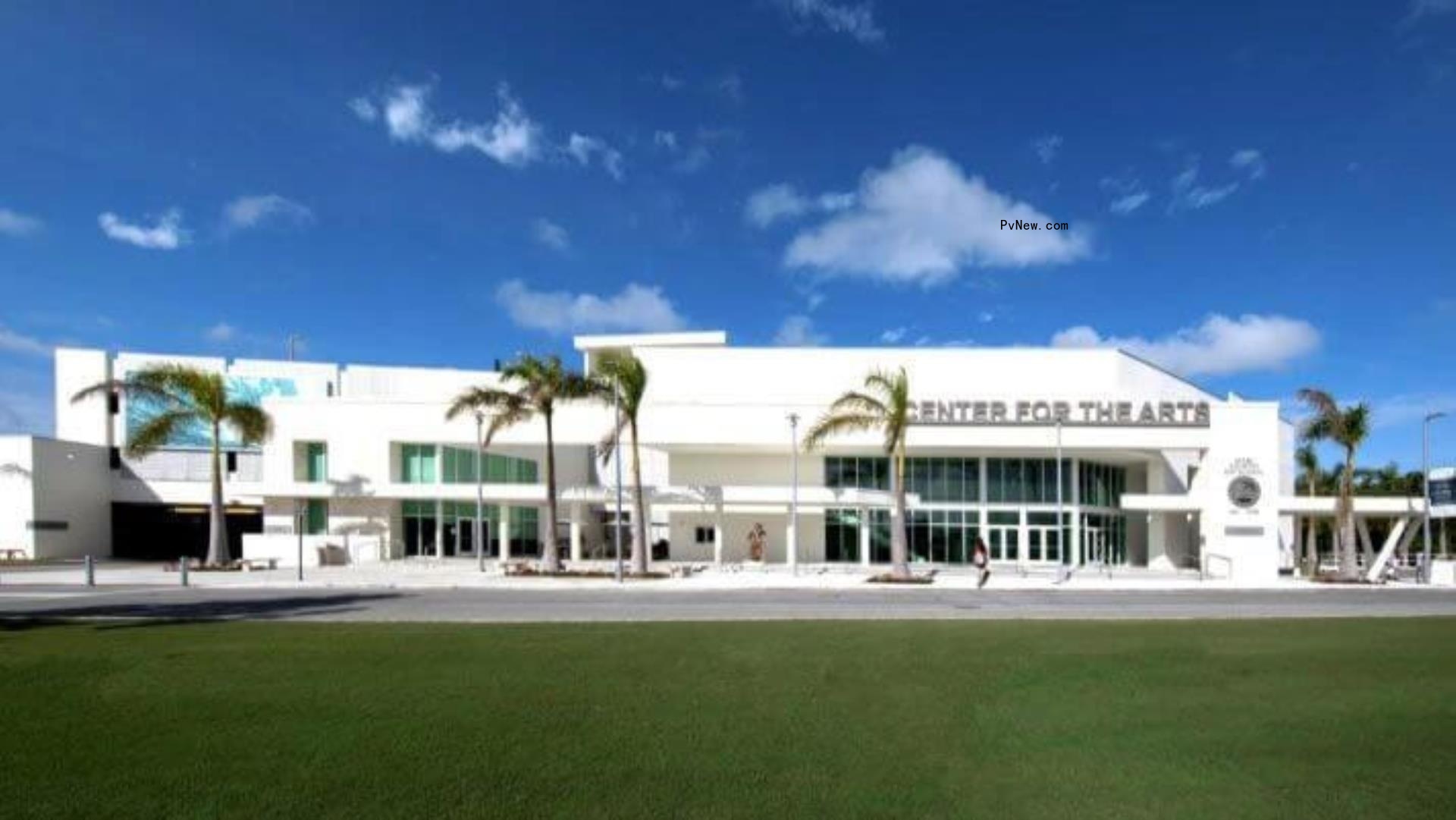 A large two-story white school building in front of a green lawn, with palm trees in front. A sign says