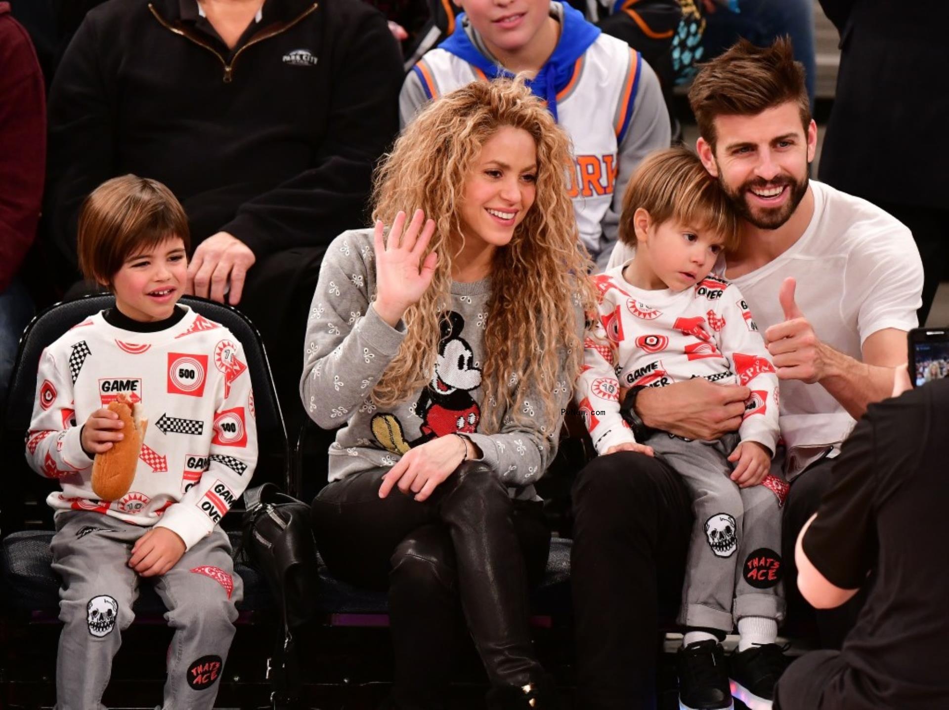 Shakira, wearing a Mickey Mouse jumper and black leather pants waves as her ex Gerard Piqué gives a thumbs up while holding his younger son on his lap. Their older son sits to their left.