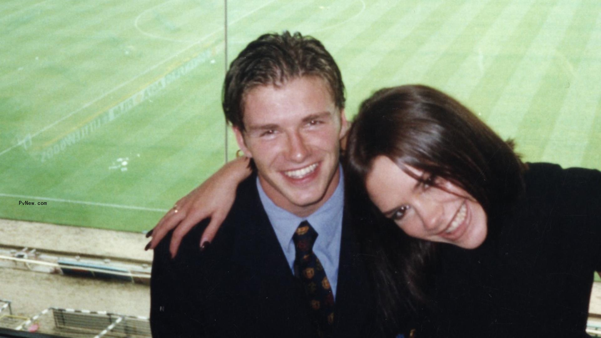 Victoria Adams cuddles David Beckham with a soccer pitch visible behind them