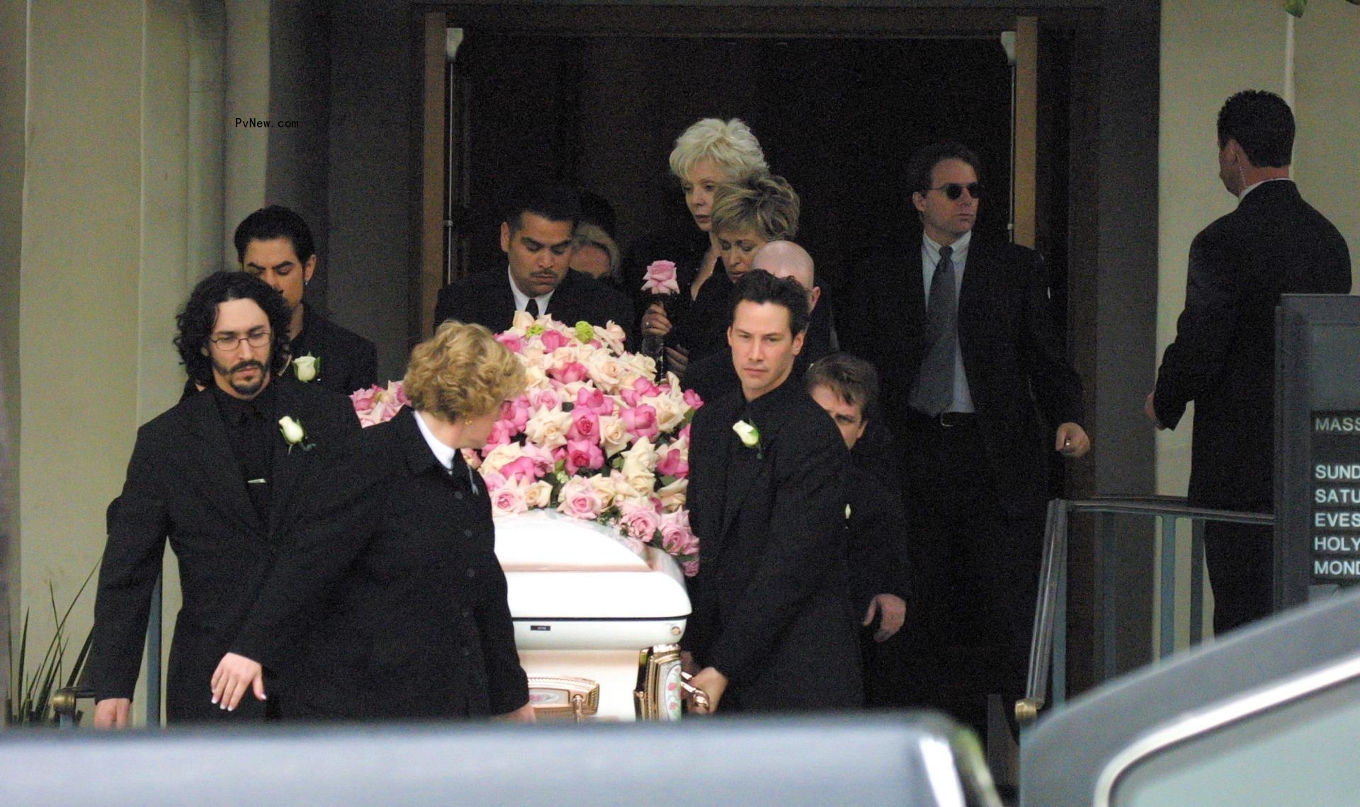Keanu Reeves holds a white coffin with pink flowers at the funeral for Jennifer Syme. Various others help with the coffin.