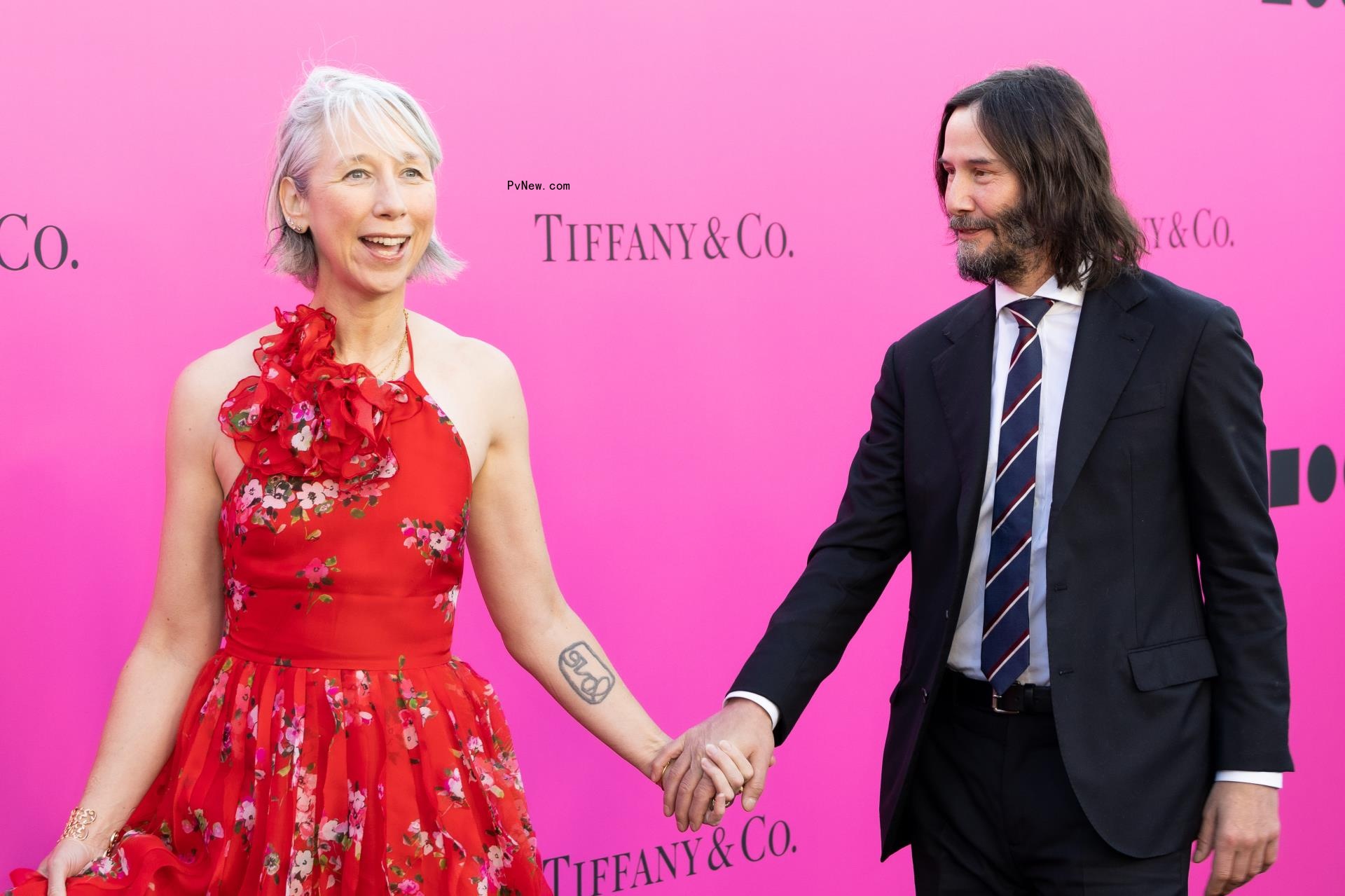 Alexandra Grant and Keanu Reeves hold hands on a red carpet, with pink Tiffany & Co. step and repeat.