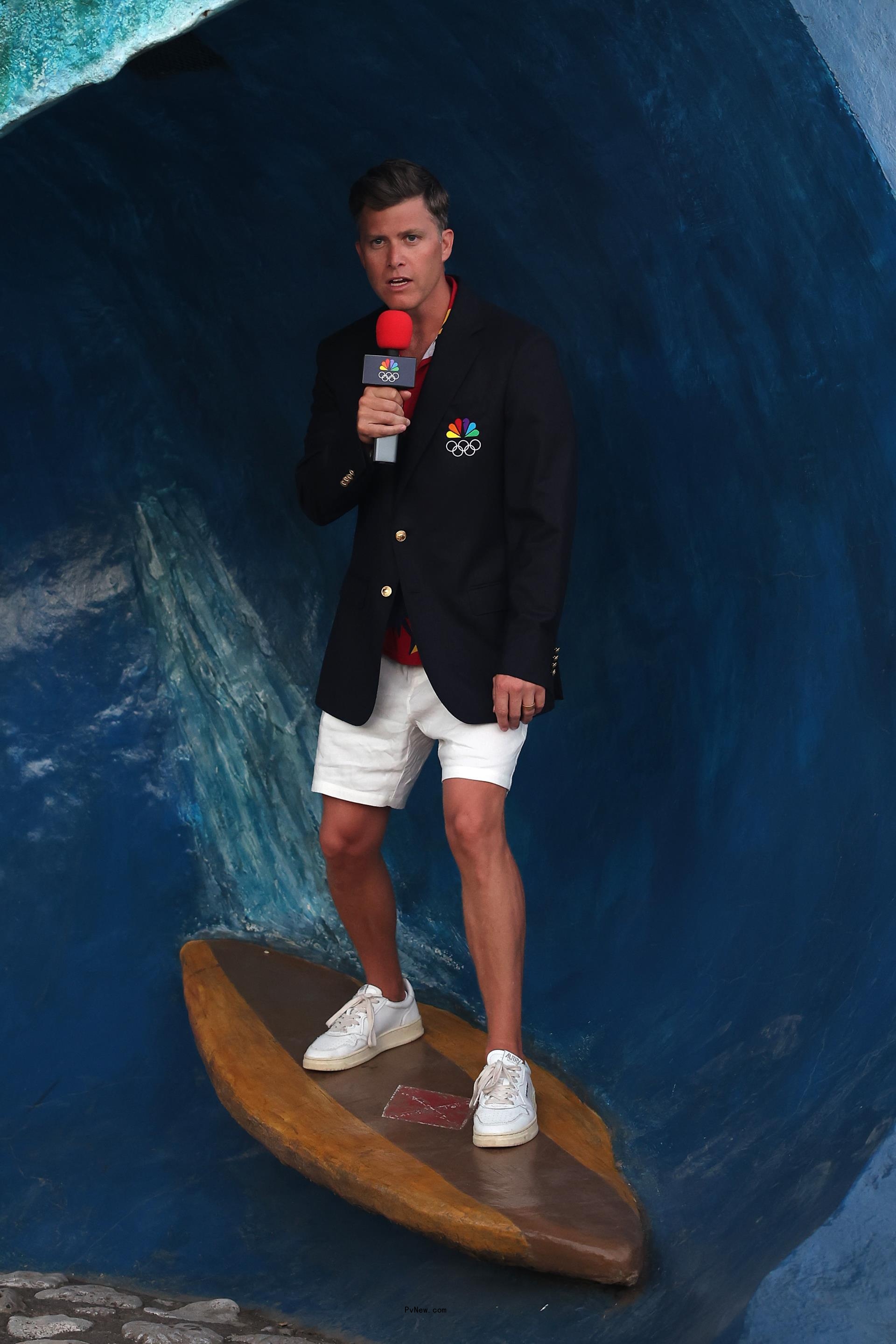 colin jost standing on a wooden surf board on a fake wave